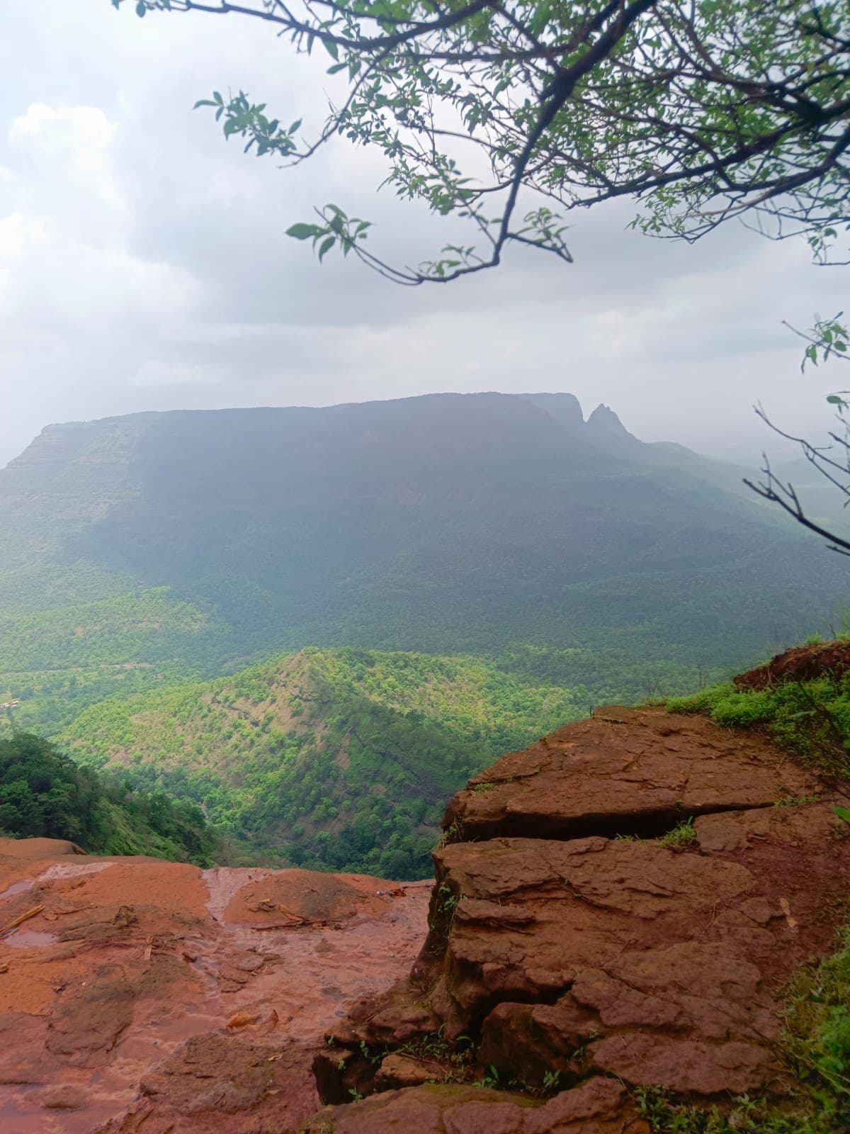 Layered red laterite rock in the foreground with a deep green valley and distant ridge behind.